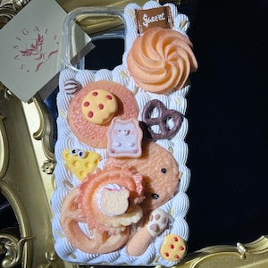 May include: A white phone case with a variety of colorful 3D decorations, including a swirl-shaped cookie, a pretzel, a donut, and a small bear. The case also features the word "Sweet" in pink lettering.