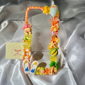 Puede incluir: Una funda transparente para teléfono con un tema de verano colorido. La funda está decorada con varios amuletos 3D, incluyendo una piña, una palmera, una estrella de mar, una magdalena y chanclas. La funda está diseñada para adaptarse a un teléfono inteligente.