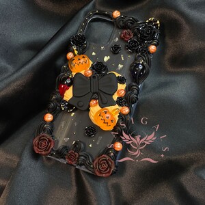 Peut inclure: Coque de téléphone noire décorée de roses noires, d'un nœud noir et de citrouilles orange. La coque a un fond noir avec des paillettes dorées et un motif noir et blanc. Le texte "Sasigals" est visible dans le coin inférieur droit.