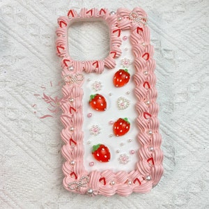 May include: A pink and white phone case with a white background and a pink border. The border is decorated with white pearls, pink swirls, and red strawberry designs.