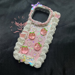 Puede incluir: Una funda de teléfono rosa y blanca con un diseño de glaseado y caramelos en forma de melocotón. La funda tiene una parte posterior transparente y un borde blanco con detalles de perlas. También hay lazos y mariposas iridiscentes en la funda.