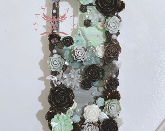 Op de afbeelding: Een zwarte telefoonhoes versierd met zwarte en witte bloemen, groene vetplanten en parels. De hoes heeft een transparant achterpaneel.