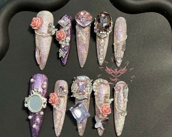 May include: A set of 12 long, pointed, false nails with various embellishments. The nails are decorated with rhinestones, pearls, flowers, and butterflies. Some nails are purple, while others are a light pink with glitter.