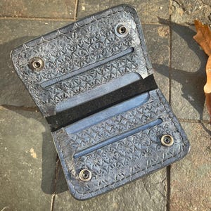 Könnte beinhalten: Eine schwarze Ledergeldbörse mit einem gemusterten Design und vier silbernen Druckknöpfen. Die Geldbörse ist aufgeklappt und zeigt zwei Kartenfächer.