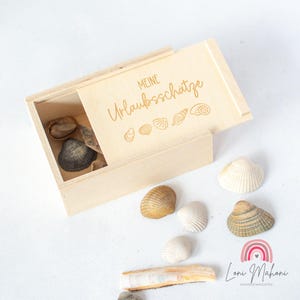 Könnte beinhalten: Eine helle Holzkiste mit Schiebedeckel, graviert mit den Worten "Meine Urlaubsschätze" und Muschelmotiven. Die Kiste ist geöffnet und zeigt Muscheln im Inneren. Mehrere Muscheln sind auf einer weißen Oberfläche verstreut.