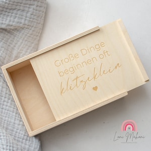 Könnte beinhalten: Eine Holzkiste mit einem Schiebedeckel. Der Deckel ist mit dem Text "Große Dinge beginnen oft klitzeklein" in goldenen Lettern graviert. Ein kleines Herz ist ebenfalls auf dem Deckel graviert.