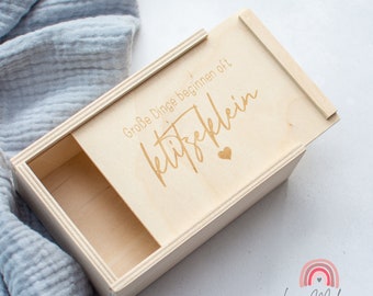 May include: A wooden box with a sliding lid. The lid has the text "Große Dinge beginnen oft  kleit". The box is made of light wood and has a natural finish.