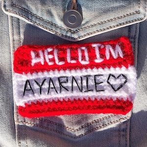 May include: A red and white crocheted name tag with the text "HELLO I'M AYARNIE" and a small heart.