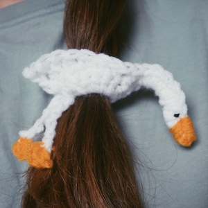 Puede incluir: Un lazo para el pelo de cisne blanco tejido a crochet con pico y patas naranjas.
