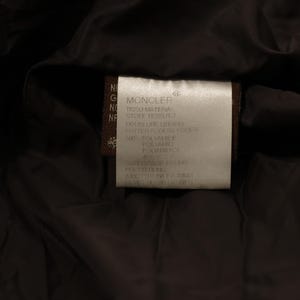 May include: Close-up of a label from a dark-colored garment. The label is rectangular, with the brand name "MONCLER" at the top. Text in multiple languages details the materials and care instructions. The background is a dark, possibly black, fabric.