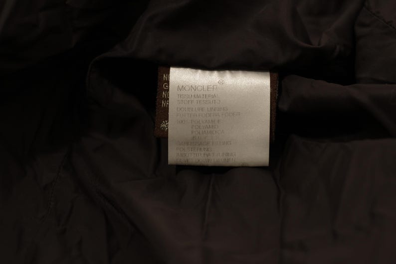 May include: Close-up of a label from a dark-colored garment. The label is rectangular, with the brand name "MONCLER" at the top. Text in multiple languages details the materials and care instructions. The background is a dark, possibly black, fabric.
