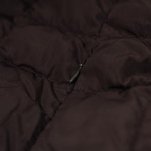 May include: Close-up of a dark brown quilted fabric, possibly a jacket or coat. The material has a subtle sheen and a diamond pattern. A silver zipper is partially visible, adding a contrasting metallic detail to the garment.