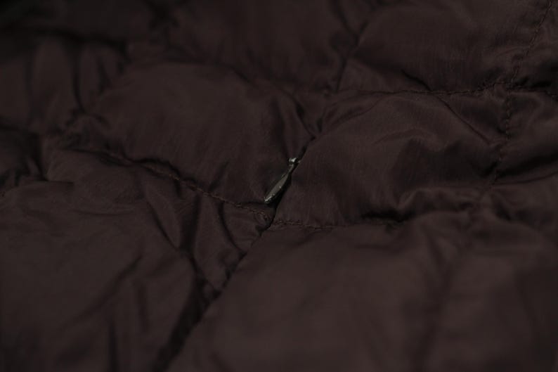 May include: Close-up of a dark brown quilted fabric, possibly a jacket or coat. The material has a subtle sheen and a diamond pattern. A silver zipper is partially visible, adding a contrasting metallic detail to the garment.
