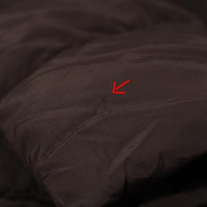 May include: Close-up of a dark brown, quilted fabric, possibly from a jacket or coat. The material has a slightly shiny appearance and a smooth texture. The image highlights the details of the stitching and the fabric's folds.