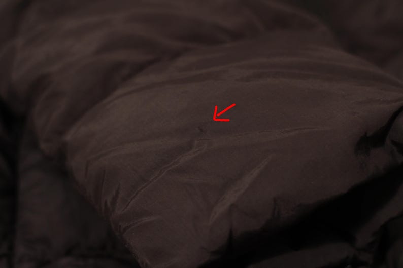 May include: Close-up of a dark brown, quilted fabric, possibly from a jacket or coat. The material has a slightly shiny appearance and a smooth texture. The image highlights the details of the stitching and the fabric's folds.