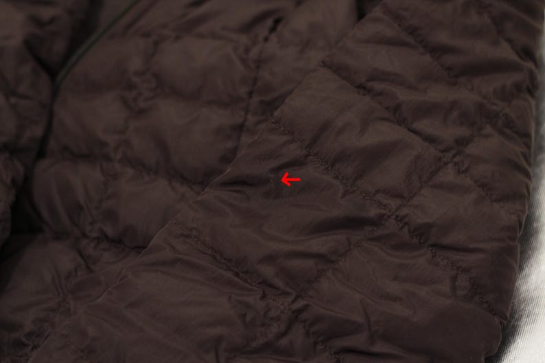 May include: Close-up of a dark brown quilted jacket. The jacket has a diamond pattern and appears to be made of a lightweight material. The fabric has a slight sheen, suggesting a possible water-resistant finish. The overall aesthetic is simple and functional.