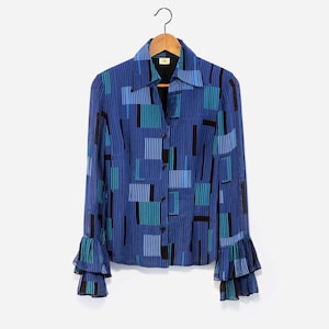 Può includere: Una camicia a maniche lunghe con motivo geometrico blu, verde e nero, con chiusura a bottoni e polsini a volant.