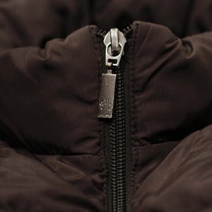 May include: Close-up of a dark brown jacket with a black zipper. The zipper pull is silver and rectangular, with a small design. The jacket's fabric appears quilted and padded.