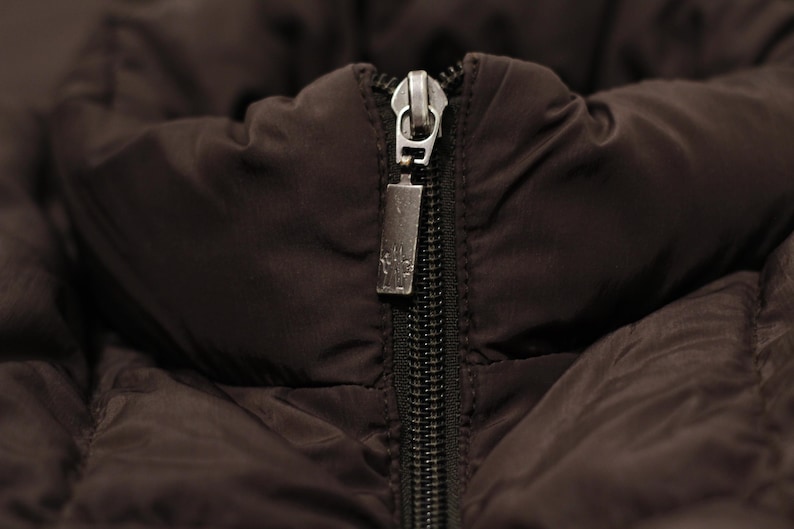 May include: Close-up of a dark brown jacket with a black zipper. The zipper pull is silver and rectangular, with a small design. The jacket's fabric appears quilted and padded.