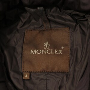 May include: Close-up of a brown label with the brand name "MONCLER" in silver lettering. The label is stitched onto a dark brown, quilted fabric. The number "3" is visible on the label.