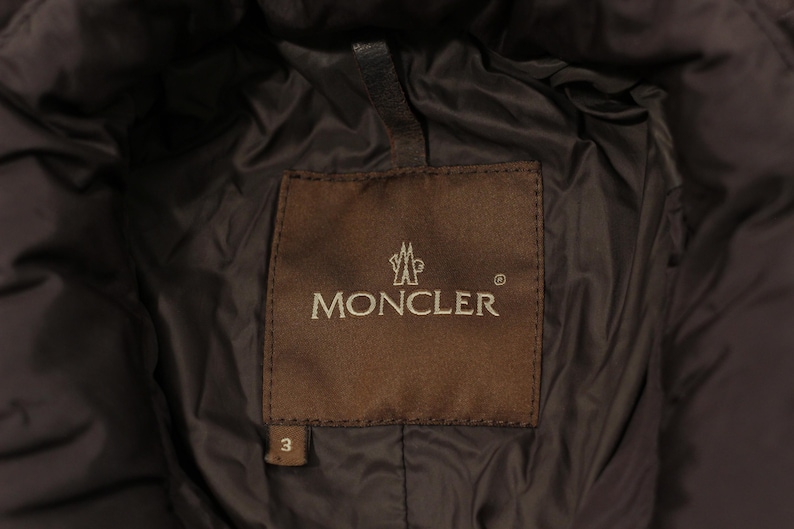May include: Close-up of a brown label with the brand name "MONCLER" in silver lettering. The label is stitched onto a dark brown, quilted fabric. The number "3" is visible on the label.