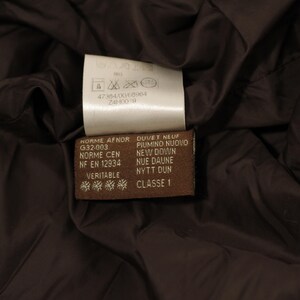 May include: Close-up of a brown garment interior with two tags. One tag is white with care symbols and text. The other tag is brown with text in multiple languages, including "DUVET NEUF" and "CLASSE 1".