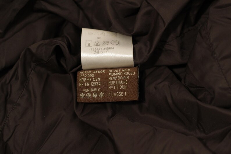 May include: Close-up of a brown garment interior with two tags. One tag is white with care symbols and text. The other tag is brown with text in multiple languages, including "DUVET NEUF" and "CLASSE 1".