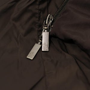 May include: Close-up of a dark brown jacket with a silver zipper. The zipper has two pulls with a logo on each. The jacket material is smooth and slightly shiny.