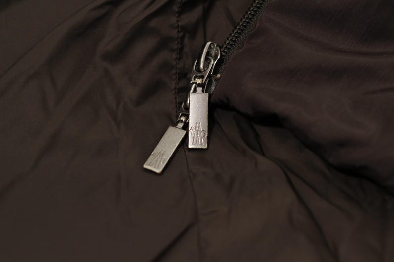 May include: Close-up of a dark brown jacket with a silver zipper. The zipper has two pulls with a logo on each. The jacket material is smooth and slightly shiny.