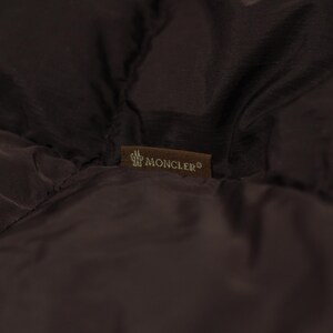 May include: Close-up of a dark brown quilted fabric with a small rectangular label. The label features the brand name "MONCLER" in white lettering and a logo. The fabric appears to be part of a jacket or coat.