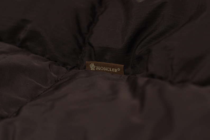 May include: Close-up of a dark brown quilted fabric with a small rectangular label. The label features the brand name "MONCLER" in white lettering and a logo. The fabric appears to be part of a jacket or coat.