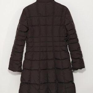 May include: A dark brown, long quilted coat hanging on a wooden hanger. The coat has a high collar and long sleeves. The coat is made of a quilted material and has a straight silhouette. The coat is a solid color and appears to be made for cold weather.