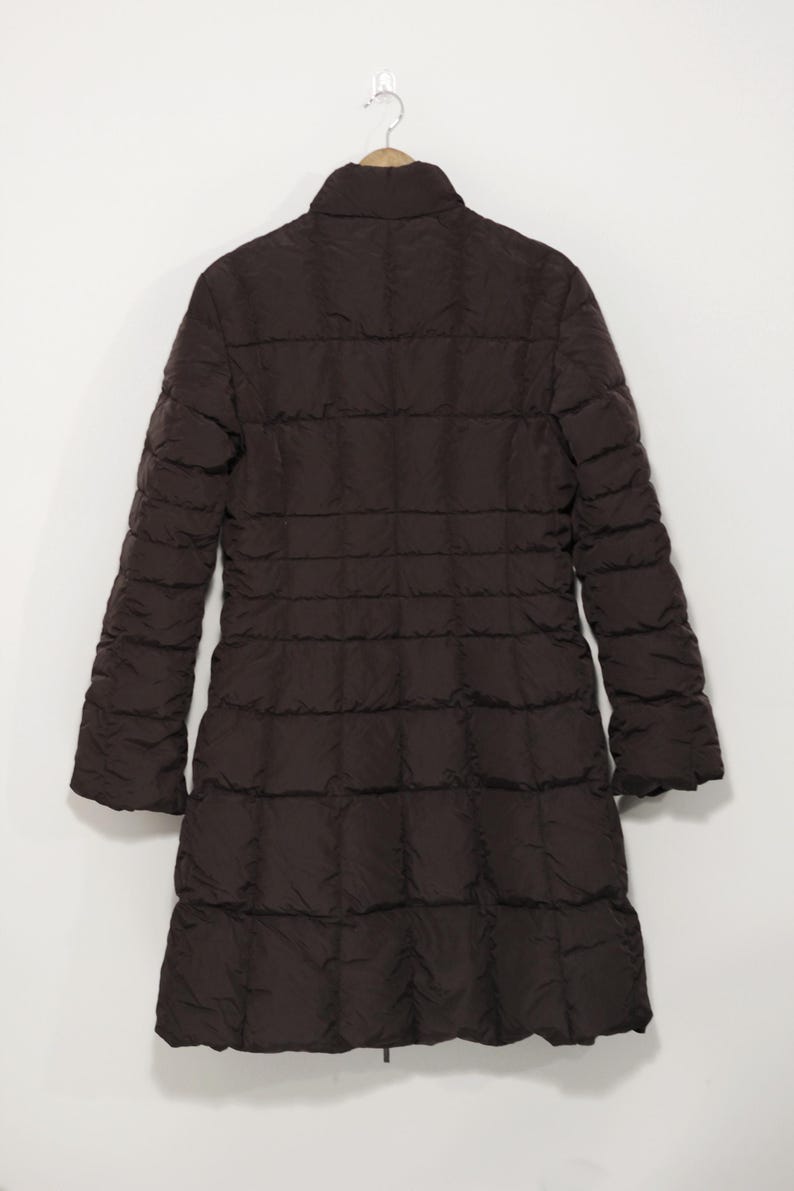 May include: A dark brown, long quilted coat hanging on a wooden hanger. The coat has a high collar and long sleeves. The coat is made of a quilted material and has a straight silhouette. The coat is a solid color and appears to be made for cold weather.