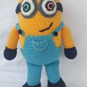 Puede incluir: Un juguete de peluche de crochet de un personaje de minion. El juguete es amarillo con un mono azul y zapatos negros. Tiene ojos grandes y redondos con pupilas negras y detalles blancos y grises. Los ojos tienen un diseño verde y rosa.
