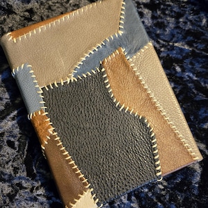 May include: A multi-colored leather journal with a patchwork design. The journal features panels of gray, blue, brown, black, and tan leather, stitched together with white thread. The cover is rectangular.