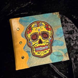 May include: A square journal with a Day of the Dead skull design. The cover features a yellow skull with red roses, blue accents, and a brown border. The journal has a teal and gold patterned background and gold corner protectors.