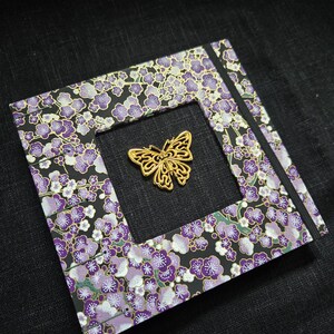 May include: A small, square, black notebook with a purple and white floral design. A gold butterfly charm is attached to the front cover.