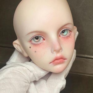 Pode incluir: Close-up do rosto de uma boneca com pele clara, l&aacute;bios fechados e olhos azuis. A boneca tem pequenas estrelas pretas tatuadas na bochecha.