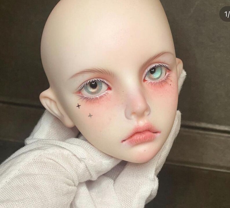 Pode incluir: Close-up do rosto de uma boneca com pele clara, l&aacute;bios fechados e olhos azuis. A boneca tem pequenas estrelas pretas tatuadas na bochecha.
