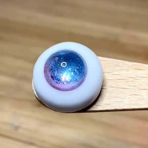 Pode incluir: Close-up de um olho de boneca azul e rosa com &iacute;ris branca e pupila preta.