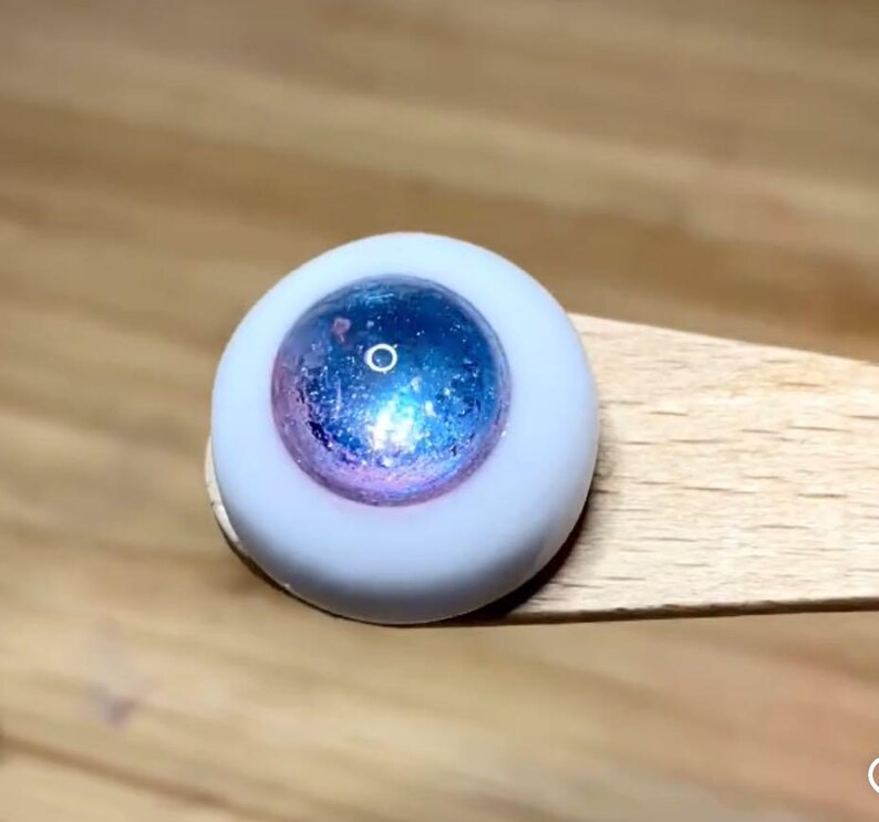 Pode incluir: Close-up de um olho de boneca azul e rosa com &iacute;ris branca e pupila preta.