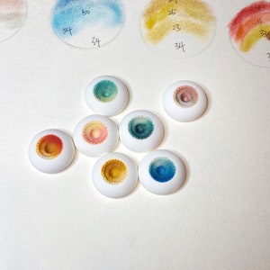 Pode incluir: Sete pequenos olhos de boneca de cer&acirc;mica branca e redondos, com &iacute;ris de cores diferentes. As &iacute;ris s&atilde;o pintadas em tons de azul, verde, amarelo e laranja.