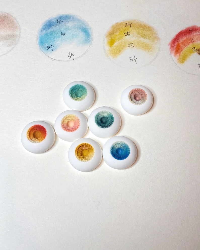 Pode incluir: Sete pequenos olhos de boneca de cer&acirc;mica branca e redondos, com &iacute;ris de cores diferentes. As &iacute;ris s&atilde;o pintadas em tons de azul, verde, amarelo e laranja.