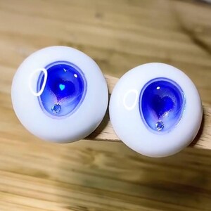 Pode incluir: Dois olhos de boneca brancos com &iacute;ris azuis e roxas e um pequeno cora&ccedil;&atilde;o prateado brilhante no centro. Os olhos t&ecirc;m uma borda branca ao redor da &iacute;ris.