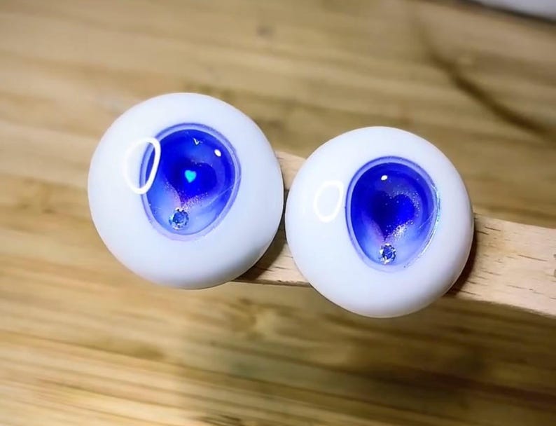 Pode incluir: Dois olhos de boneca brancos com &iacute;ris azuis e roxas e um pequeno cora&ccedil;&atilde;o prateado brilhante no centro. Os olhos t&ecirc;m uma borda branca ao redor da &iacute;ris.