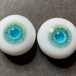 Pode incluir: Um par de olhos de vidro azul e verde com esclera branca. As &iacute;ris s&atilde;o azuis claras com um anel externo verde e um anel interno amarelo.