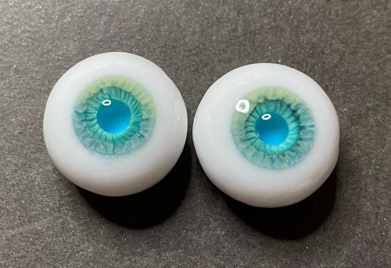 Pode incluir: Um par de olhos de vidro azul e verde com esclera branca. As &iacute;ris s&atilde;o azuis claras com um anel externo verde e um anel interno amarelo.