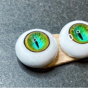 Pode incluir: Dois olhos de boneca brancos com &iacute;ris verdes e azuis e pupilas pretas. Os olhos est&atilde;o sobre uma superf&iacute;cie de madeira.