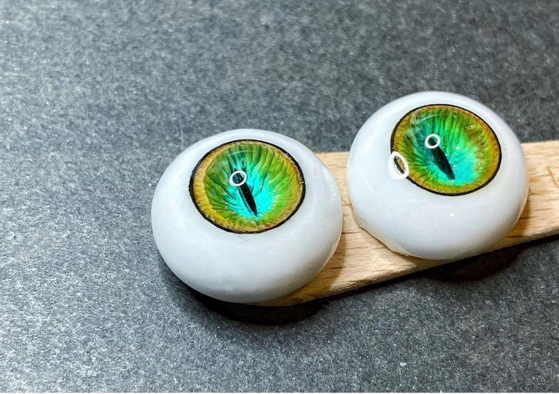 Pode incluir: Dois olhos de boneca brancos com &iacute;ris verdes e azuis e pupilas pretas. Os olhos est&atilde;o sobre uma superf&iacute;cie de madeira.