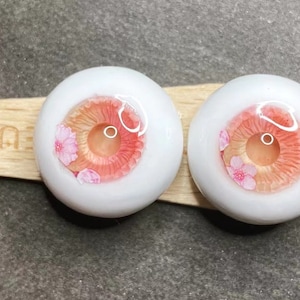 Pode incluir: Dois olhos de boneca brancos com &iacute;ris rosa e p&ecirc;ssego e um design floral. Os olhos est&atilde;o sobre uma superf&iacute;cie de madeira.